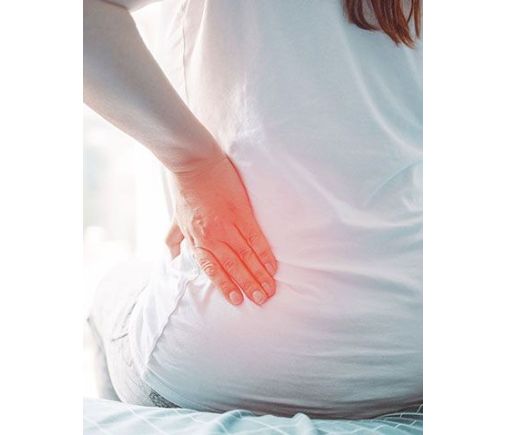 Woman sitting on bed, feeling her lower back, which is marked with red to indicate pain.